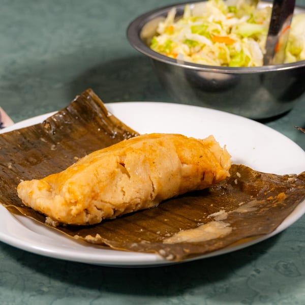 Golden tamale on a banana leaf, served on a white plate with salad and red sauce.