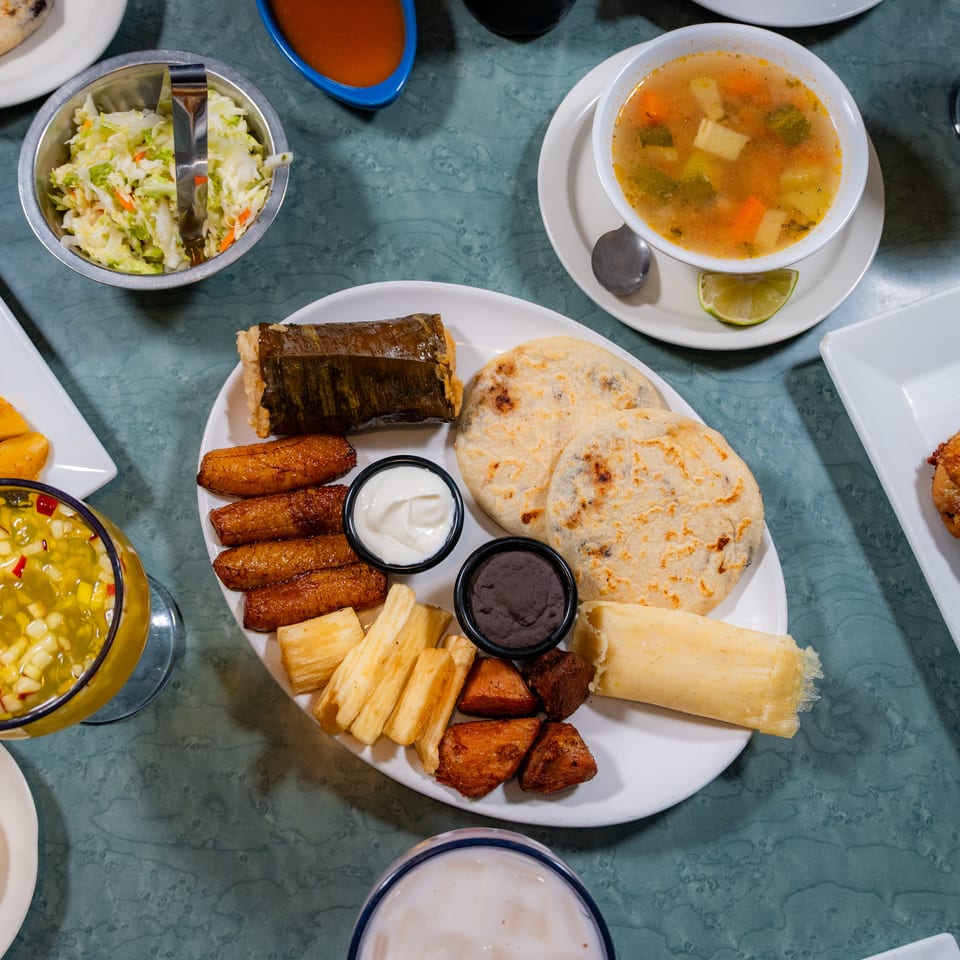 Restaurant table with a spread of Latin American food: pupusas, chicken, plantains, soup, and tamales.