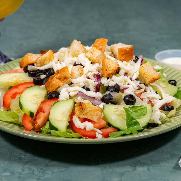 Green plate of fresh salad with lettuce, tomatoes, cucumbers, olives, shredded cheese, and croutons.